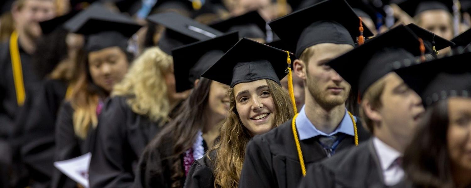 SPU graduates line up to begin the Commencement ceremony; one young graduate leans to her left, 微笑, to look toward the front of the stage.